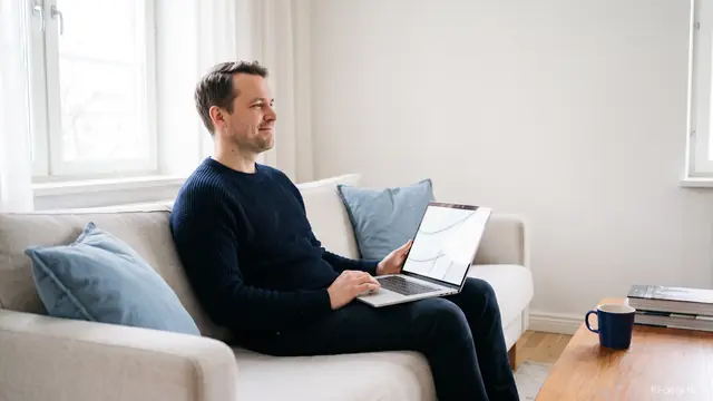 Mann sitzt mit einem Laptop auf dem Sofa neben einem hellen Fenster